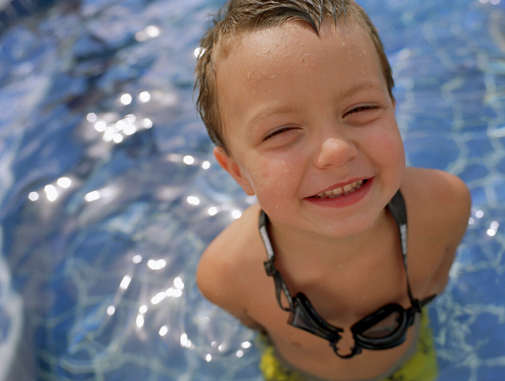 Can I Teach My Kids to Swim with a Swim Spa? 3 young boy standing in a swim spa or similar product. He is looking up at the camera, smiling, with goggles around his neck.