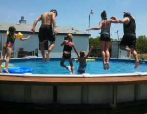 Pictured is a family of six who are all mid-air about to jump into their above-ground pool. The family is facing away from the camera and they are jumping off of a wooden ledge into a pool that is ahead of them.