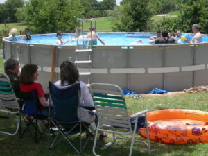 An above ground swimming pool is pictured with several kids playing inside while three adults sit outside of the pool in lawn chairs watching the children.
