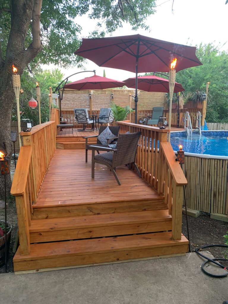 How to Rebalance Pool Water After a Pool Party 6 An above-ground pool is pictured on the right side of the image. There is a wooden deck with steps leading up to the pool. There are tiki torches, chairs, and umbrellas on the deck.