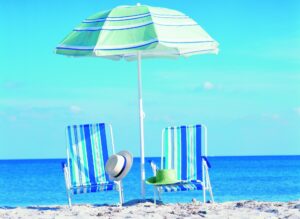 Two beach chairs are side by side on the sand in front of the ocean. There is a sunhat sitting on each chair and an umbrella covering them.