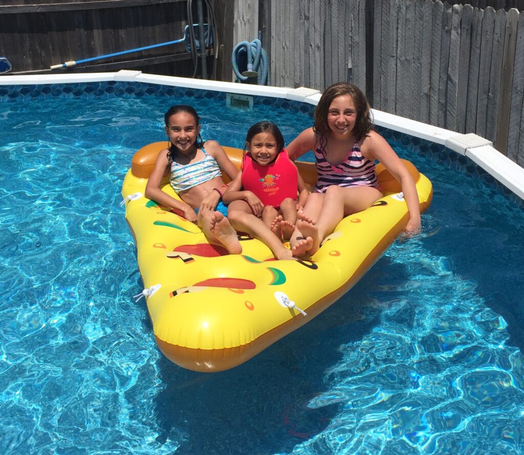 8 Best Above Ground Pool Benefits for the Active Family 7 Three kids sitting on a pizza-shaped pool float in an above ground pool.