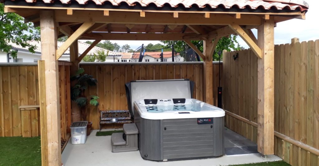 Home Considerations To Make Before Purchasing a Hot Tub 1 Alt Text: An outdoor hot tub sits beneath a wooden shelter with a red tile roof inside a fenced backyard with green grass.