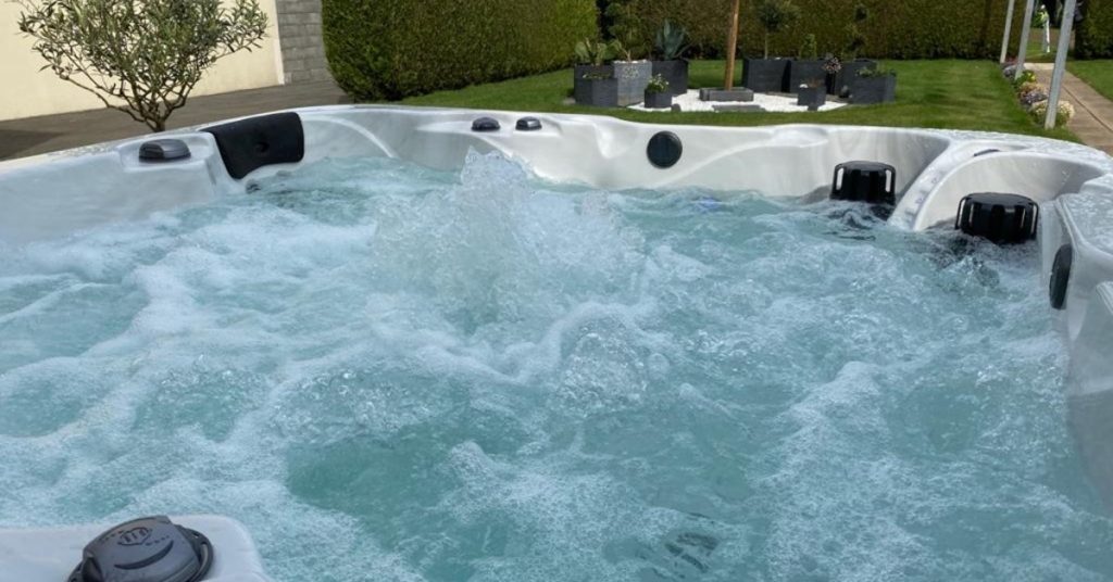 Home Considerations To Make Before Purchasing a Hot Tub 2 A bubbling outdoor hot tub has white molded seats and black headrests. A hedge and green lawn are in the background.