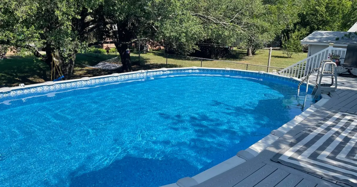 Salt vs. Chlorine Pool Systems: The Differences Explained 2 A deck with a striped rug is beside an above-ground pool with bright blue water. Tall trees are in the background.