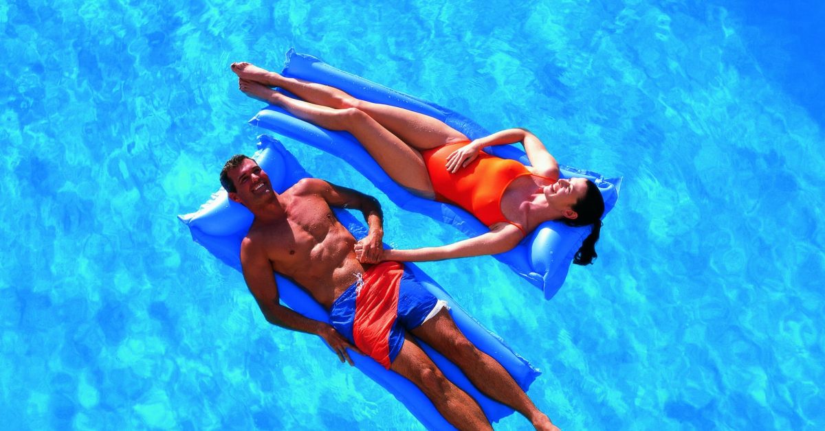 A man and a woman each laying on a floating lounge in a pool. They are opposite one another and are holding hands while smiling after their pool opening