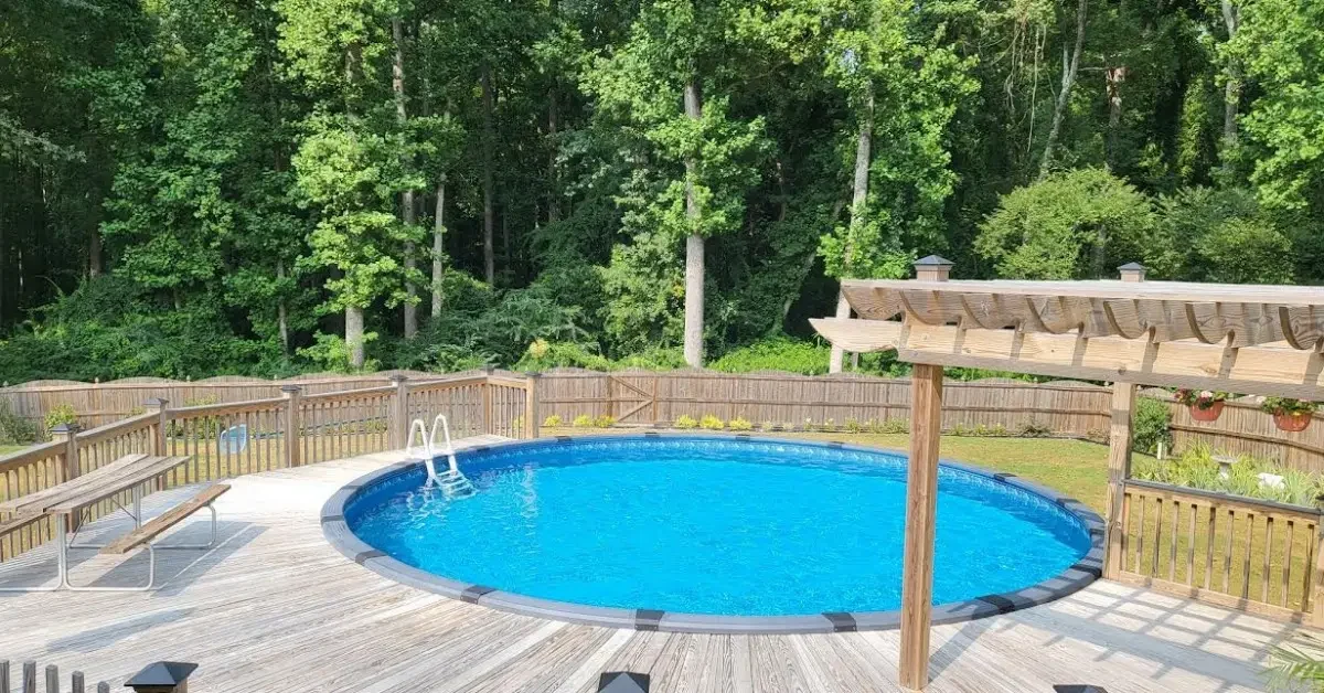 A round, above-ground pool built into a wood deck with a railing, a pergola, and benches. Trees are in the background.
