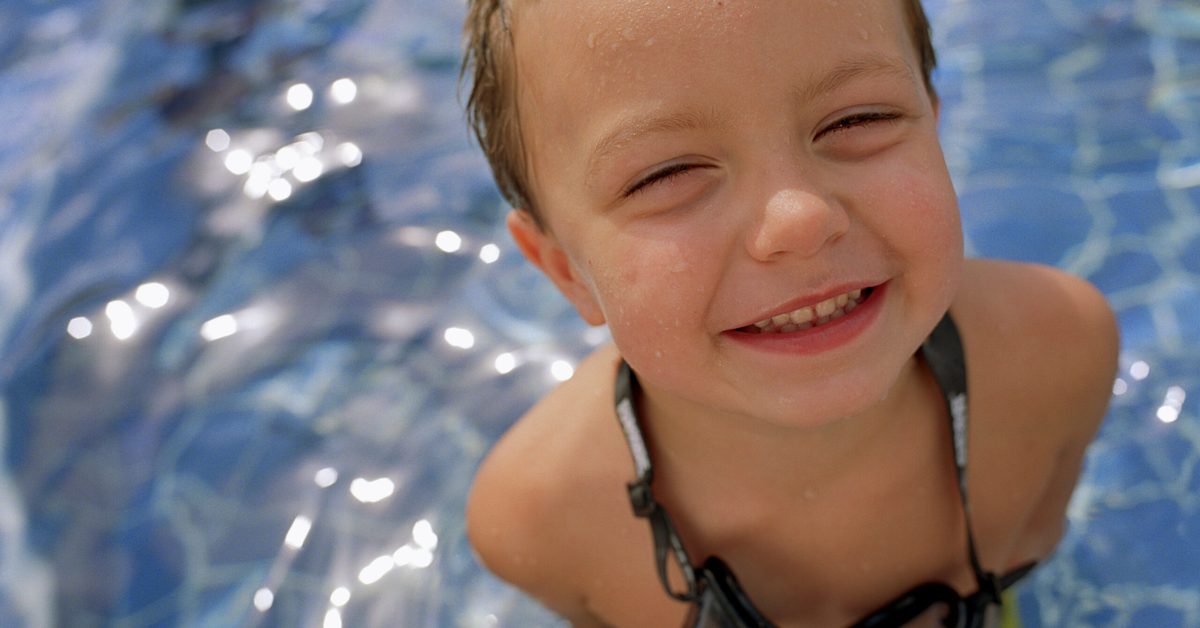 young boy standing in a swim spa or similar product. He is looking up at the camera, smiling, with goggles around his neck.
