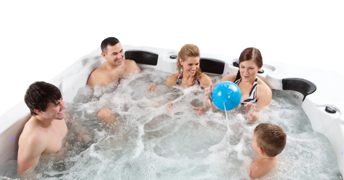 Pictured is a family of five sitting together in a swim spa. The swim spa jets are running and the family is playing catch with a blue ball.