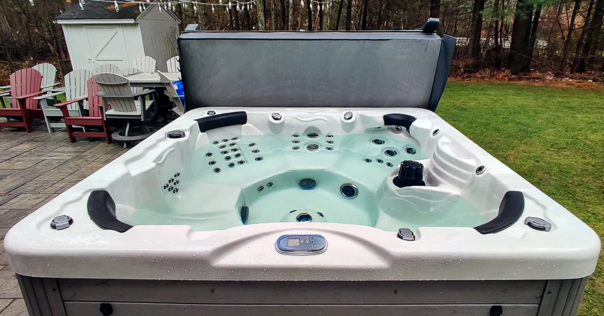 Pictured is a hot tub viewed looking down from a standing viewpoint. The jets are off to show a clear image of the inner shell. A cover can be seen behind the hot tub. There are some patio chairs and a small shed to the left of the tub, and a grassy yard to the right.